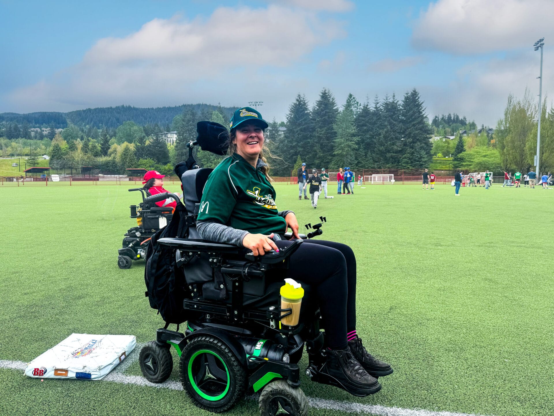 A smiling woman in a wheelchair wearing a green sports jersey and cap sits on a grassy sports pitch. Other people and trees are visible in the background under a partly cloudy sky.