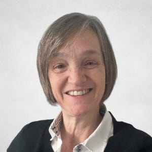 Ann Rae is a smiling older woman with short grey hair and is wearing a white collared shirt and a black jumper.