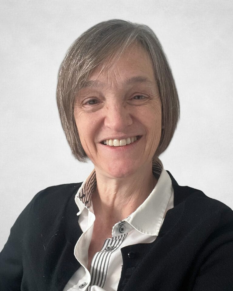Ann Rae is a smiling older woman with short grey hair and is wearing a white collared shirt and a black jumper.