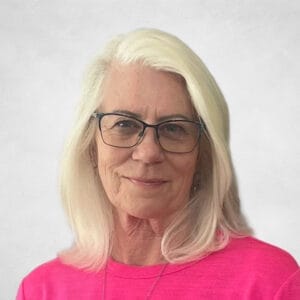 Bonnie Nybo is a woman with long white hair and glasses, wearing a bright pink top and a necklace.
