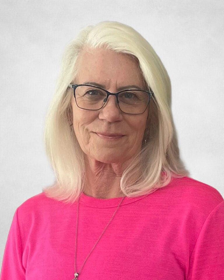 Bonnie Nybo is a woman with long white hair and glasses, wearing a bright pink top and a necklace.