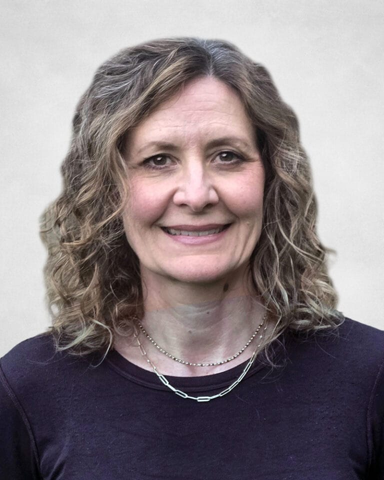 Carolyn Janzen is a woman with curly, shoulder-length light brown hair. She is wearing a dark top and two layered necklaces.