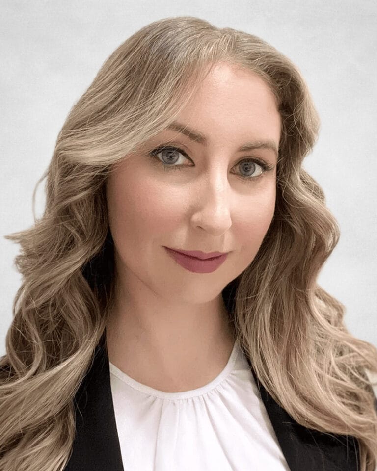 Cassandra Howlett is a woman with long, wavy blonde hair and light eyes, wearing a white blouse and black jacket.