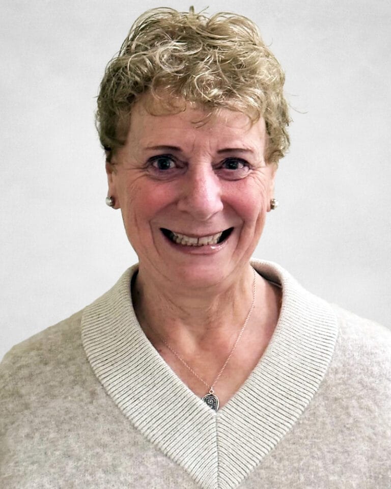 Diane Gerrard is an older woman with short, curly blonde hair. She wears a light beige jumper with a wide collar and a silver necklace.