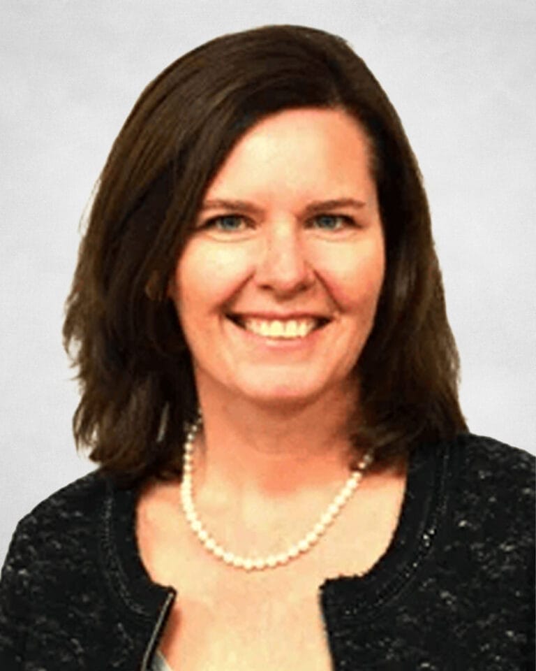 Heather McMurtry is a woman with shoulder-length brown hair, wearing a black textured jacket, a light-coloured top, and a pearl necklace.