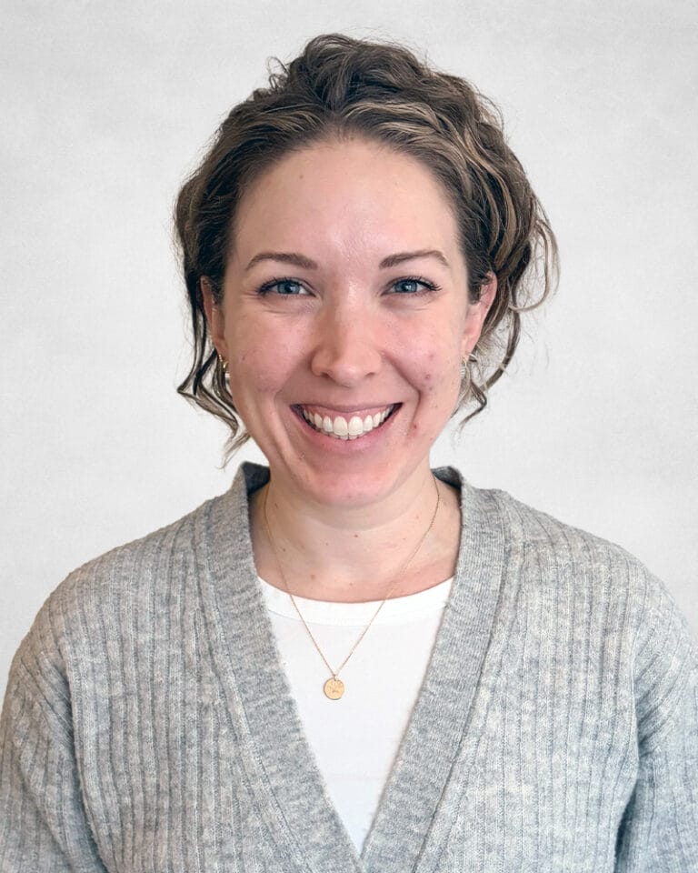 Jenna Cookie is a woman with wavy, light brown hair. She is wearing a grey cardigan over a white shirt and a gold necklace with a small circular pendant.