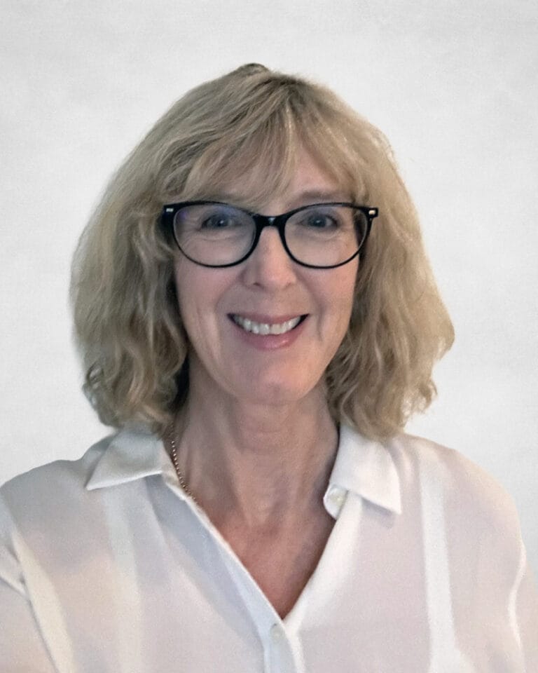 Jo Anne Chisholm is a smiling woman with shoulder-length blonde hair and glasses. She is wearing a white collared shirt.