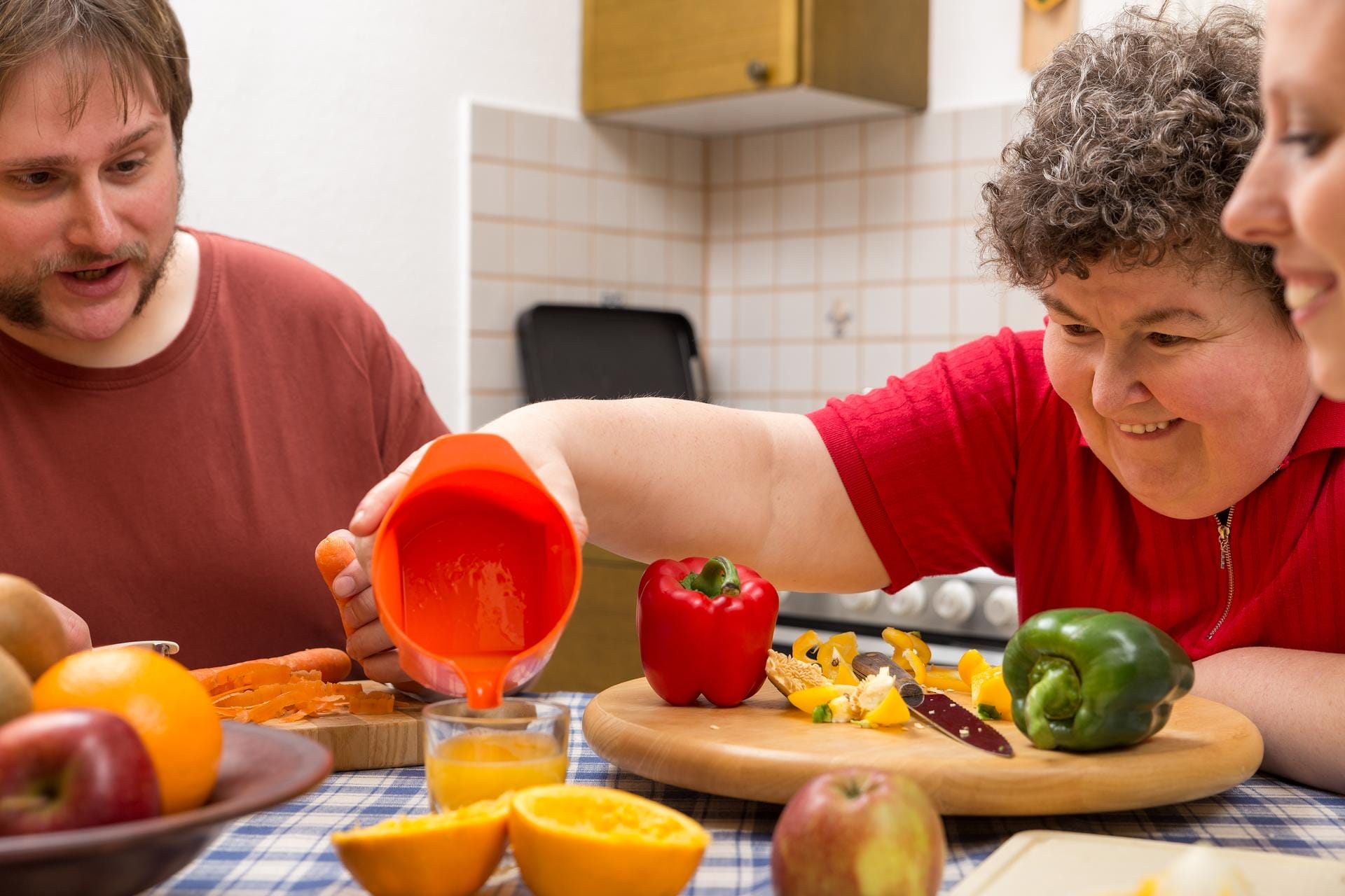 Three people are preparing food together in a kitchen. One person pours orange juice into a glass, while fresh fruit and vegetables, such as peppers and oranges, are arranged on the table. They all appear to be enjoying the activity.