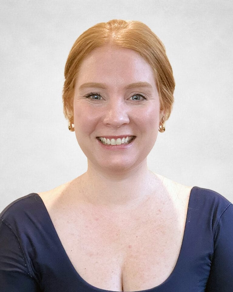 Karalyn Mayne is a woman with light skin and red hair pulled back. She is wearing gold hoop earrings and a navy blue top with a scoop neckline.