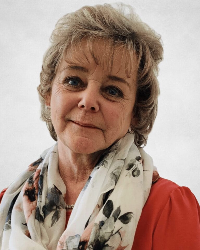 Karen Desormeau is an older woman with short, wavy, light brown hair wears a red top and a white scarf with a floral pattern.