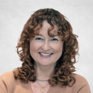 Lindsay Alford is a woman with curly brown hair and fair skin. She is wearing a light brown top and two necklaces, one with a gold pendant.