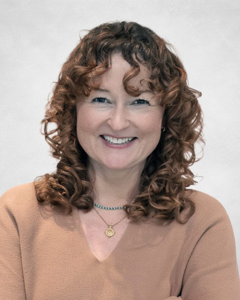 Lindsay Alford is a woman with curly brown hair and fair skin. She is wearing a light brown top and two necklaces, one with a gold pendant.
