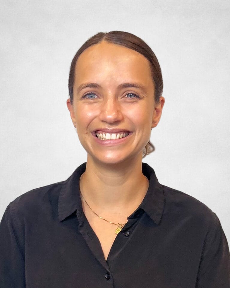 Lucy Friend is a woman with fair skin and blue eyes and has tied-back brown hair. She is wearing a black button-up shirt and a gold necklace.