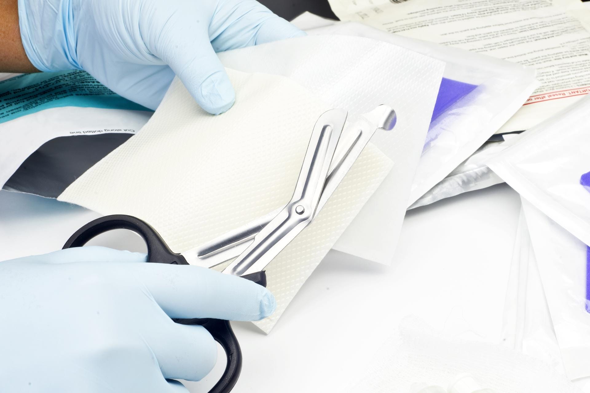 A person wearing blue medical gloves uses bandage scissors to cut a piece of white gauze, with medical supplies and packaging visible in the background.