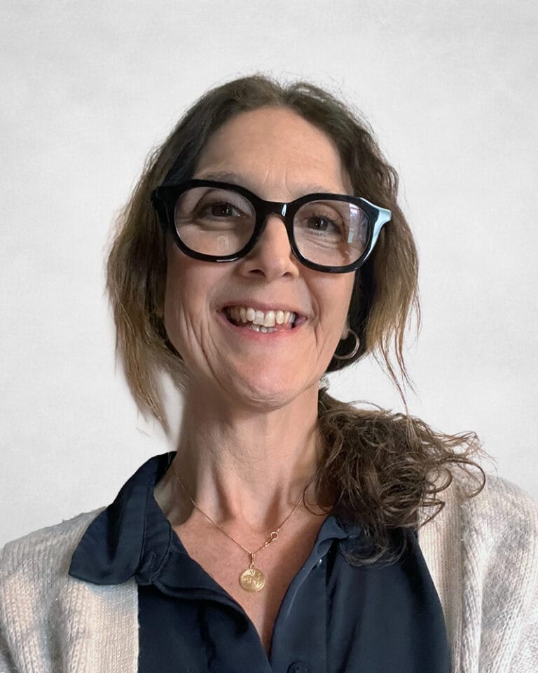 Najade Minns is a woman with curly brown hair. She is wearing large black glasses, a navy blouse, a light cardigan, and a gold necklace.