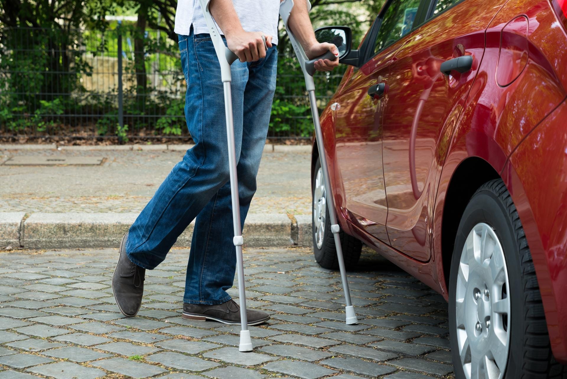 A person using crutches stands next to a red car on a cobbled street, wearing blue jeans and brown shoes. Only the lower half of the person is visible.