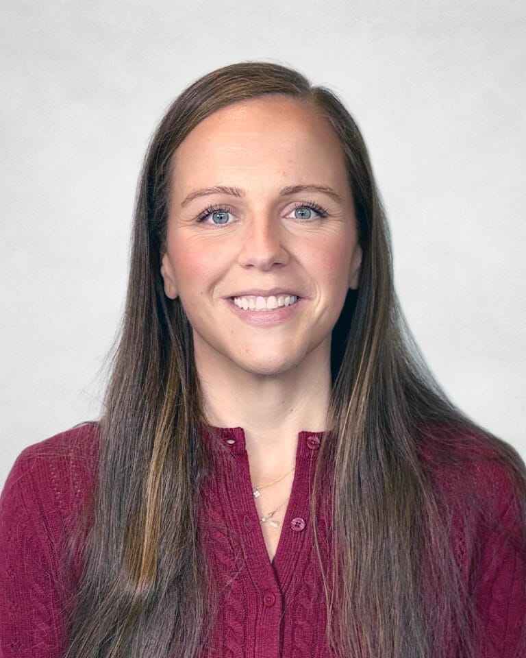 Nicole Armstrong, A woman with long brown hair, wearing a maroon knitted jumper, smiles at the camera against a light grey background.