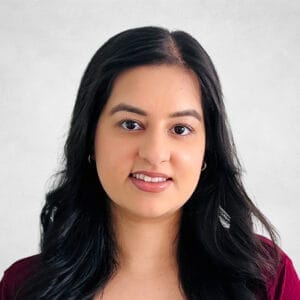 Preety Sian, A woman with long, wavy black hair and hoop earrings is smiling slightly. She is wearing a burgundy top and is posed against a light grey background.