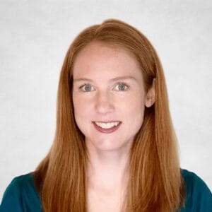 Vanessa Sturdy is a woman with long straight red hair, light skin, and green eyes. She is wearing a teal top and a necklace with a small oval pendant.