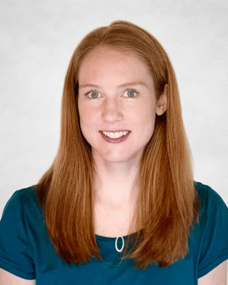 Vanessa Sturdy is a woman with long straight red hair, light skin, and green eyes. She is wearing a teal top and a necklace with a small oval pendant.