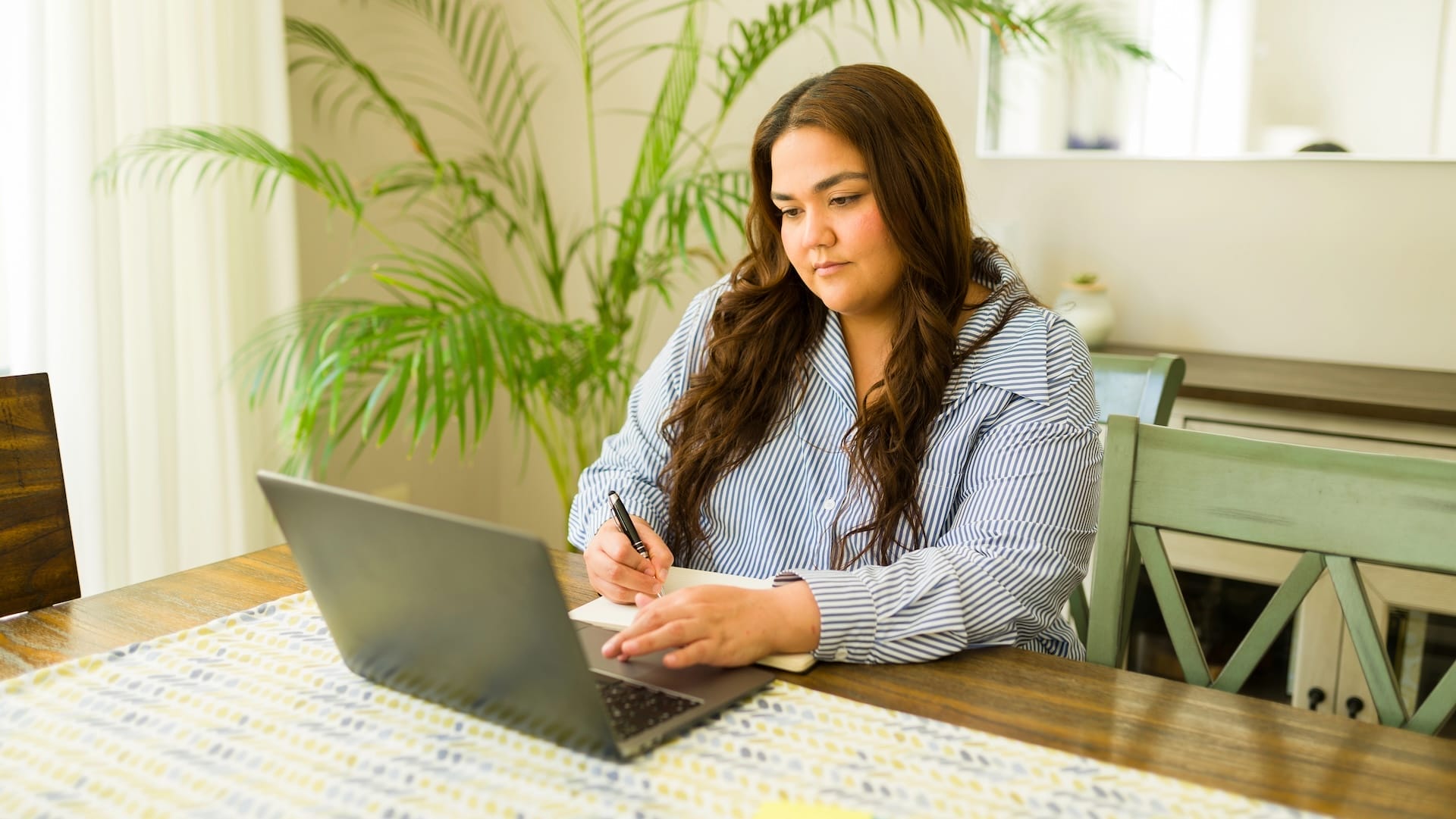 A woman with long brown hair sits at a wooden table, writing in a notebook while looking at a laptop. She is in a bright room with a green plant and natural light coming through the window.