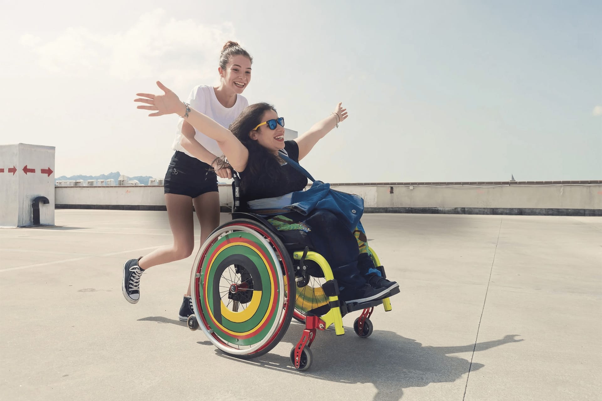 A young woman pushes a girl in a colourful wheelchair outdoors on a sunny day. The girl in the wheelchair has her arms spread wide and is smiling, while both appear joyful and carefree.