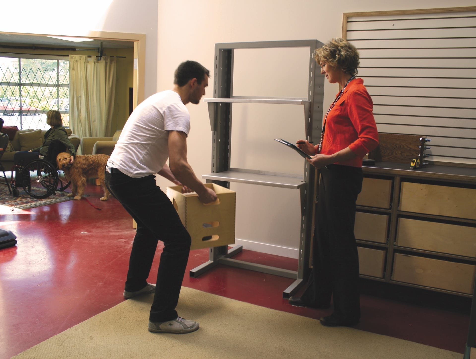 A man lifts a box using proper form while a woman with a clipboard observes.