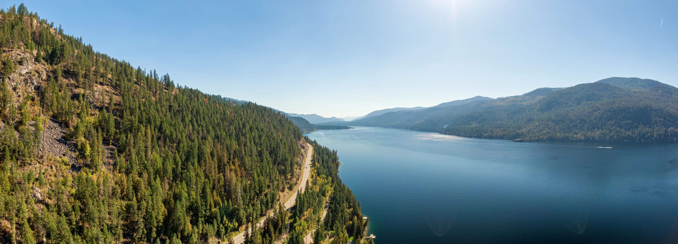 A panoramic view of a large lake bordered by forested hills and mountains under a clear blue sky, with a winding road running alongside the water’s edge.