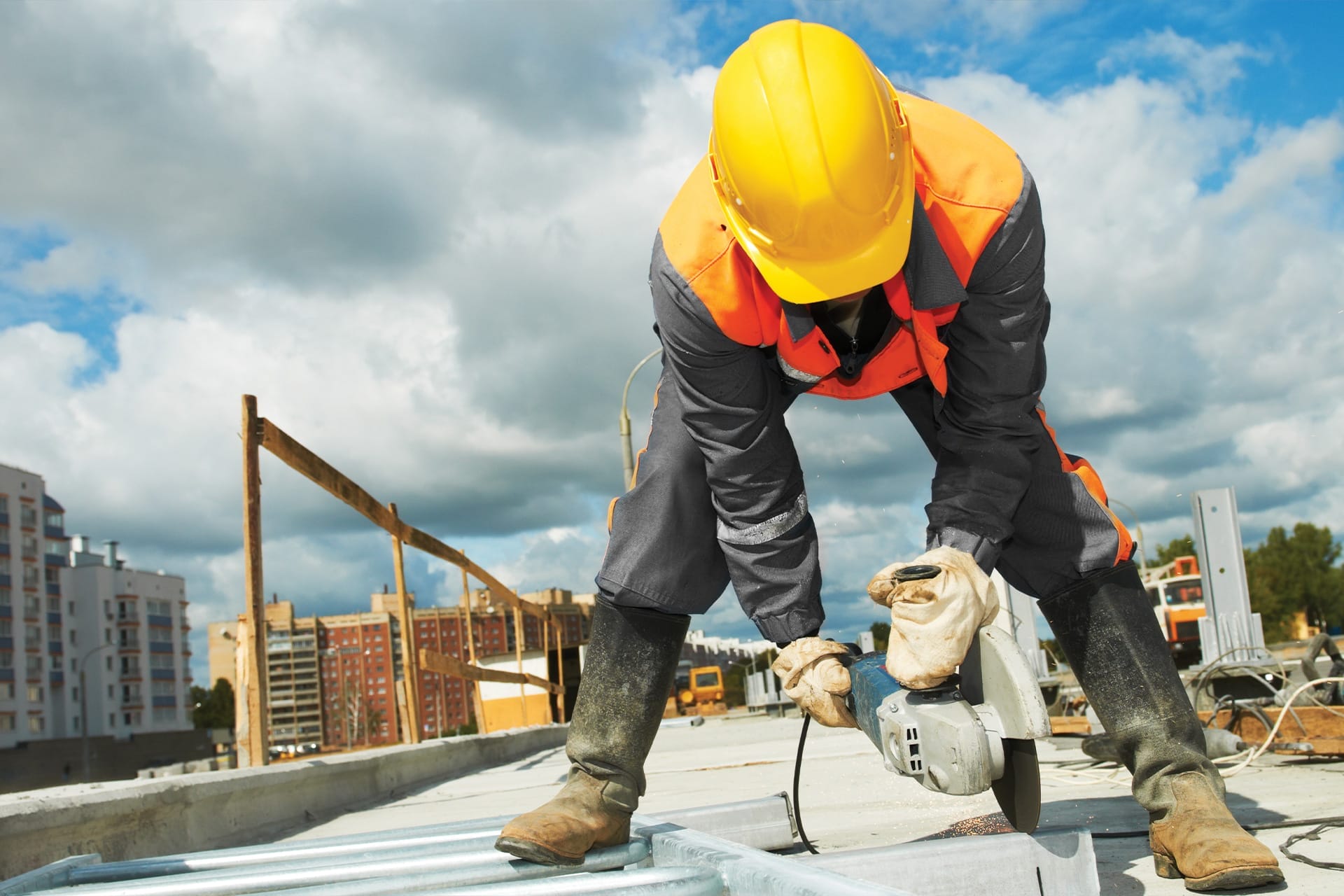 A construction worker in a yellow hard hat and orange vest uses a power tool to cut metal on a building site, with city buildings and cloudy sky in the background.