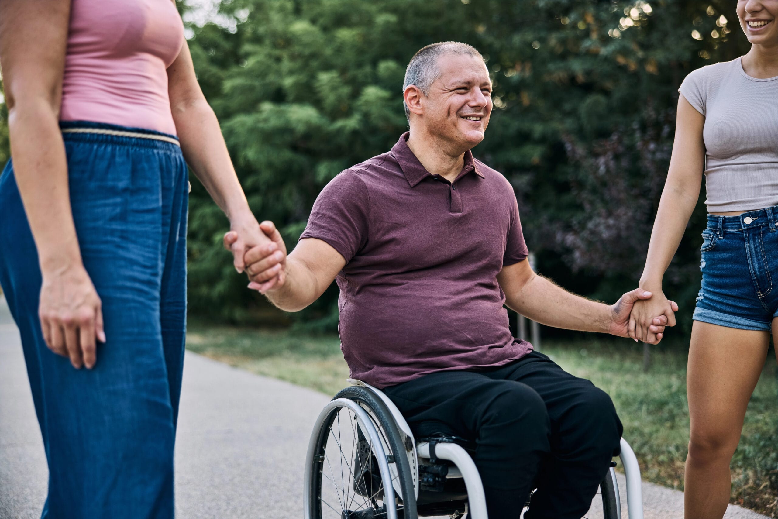 A man in a wheelchair holds hands with two people while outdoors, smiling. Trees and greenery are visible in the background.