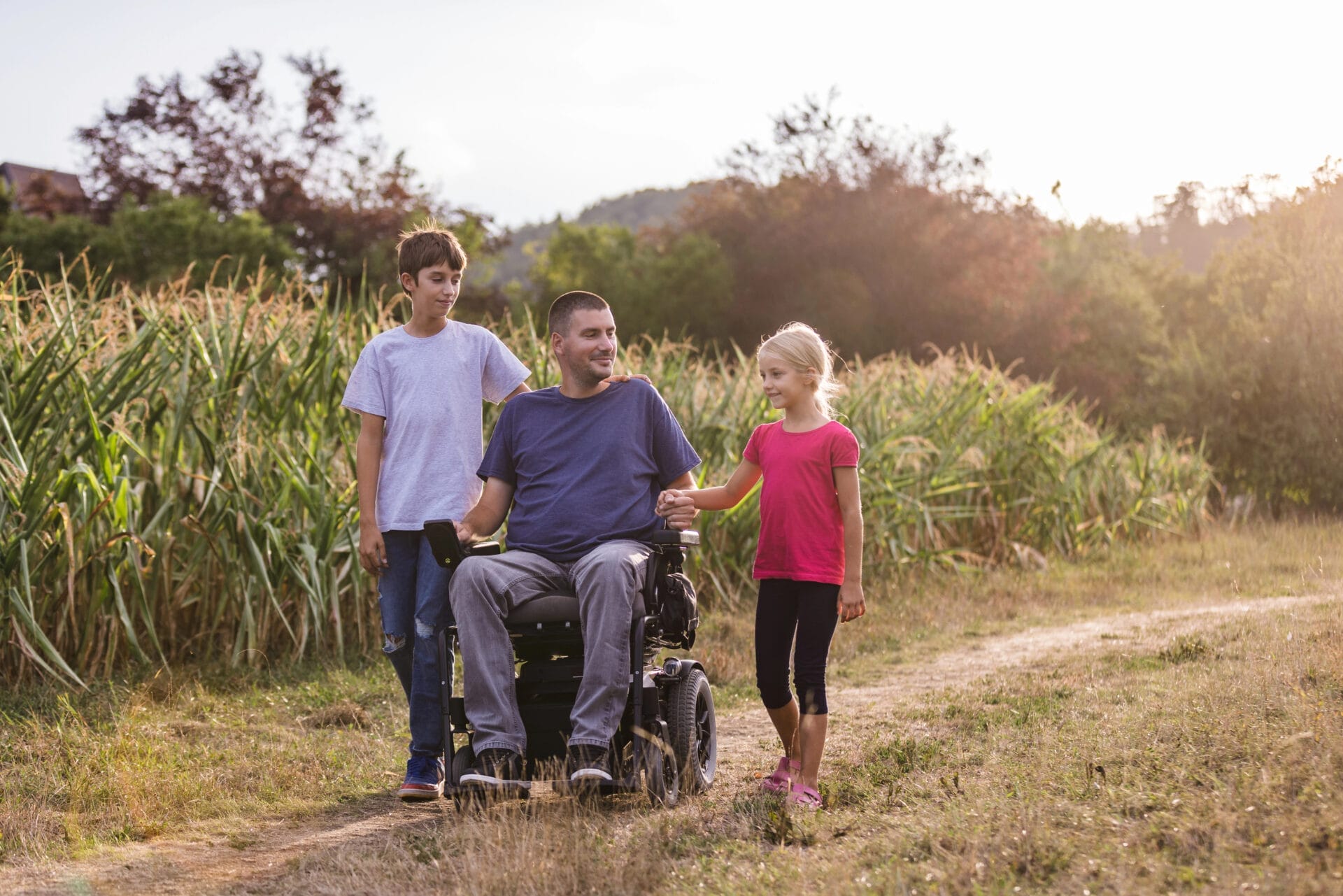 A man in a wheelchair is on a dirt path outdoors, accompanied by a young boy and a young girl. They are walking together near tall grass and trees, with sunlight in the background.