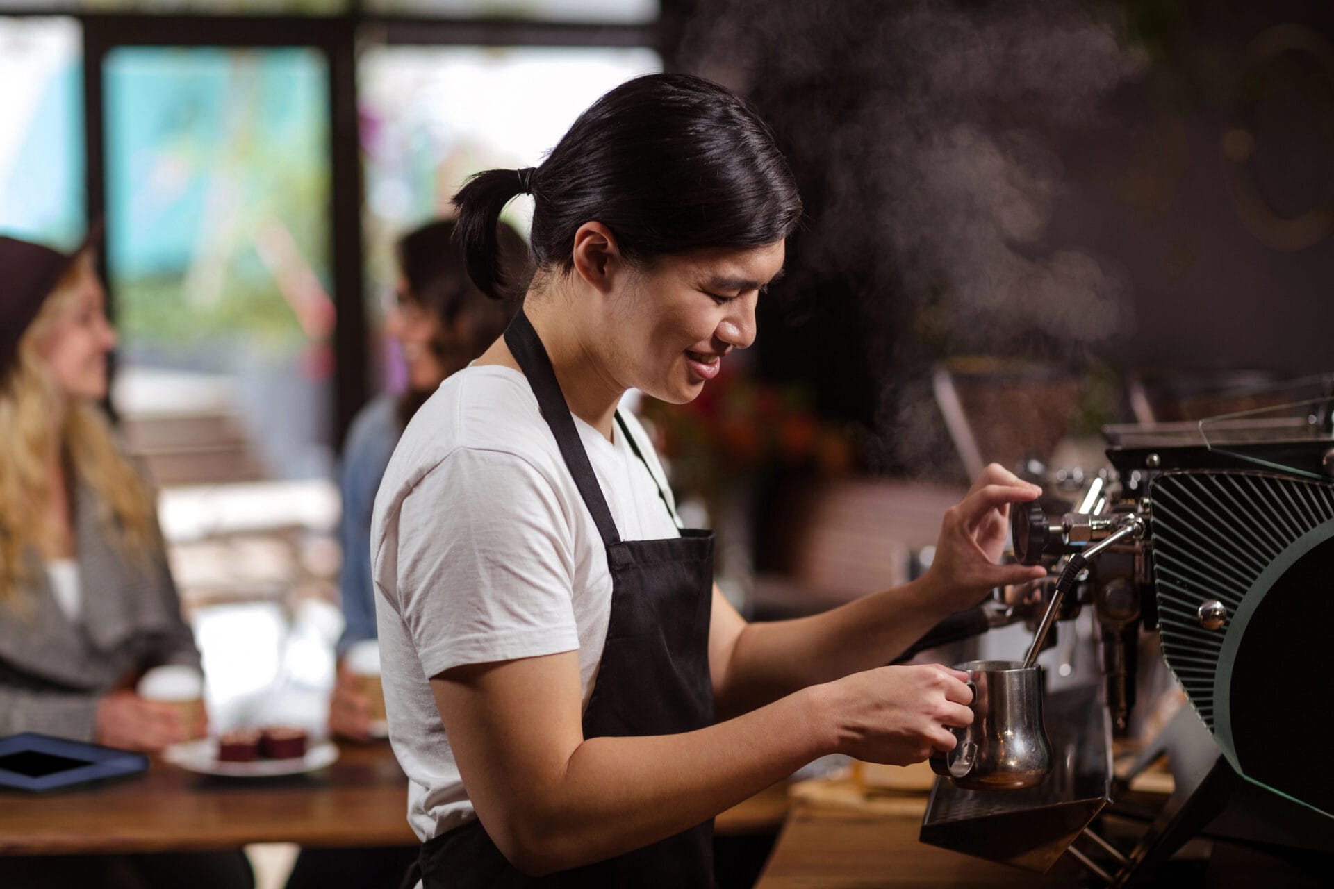 A barista wearing a black apron steams milk using an espresso machine in a café, with two customers sitting and chatting in the background. Steam rises from the jug as they focus on their task.