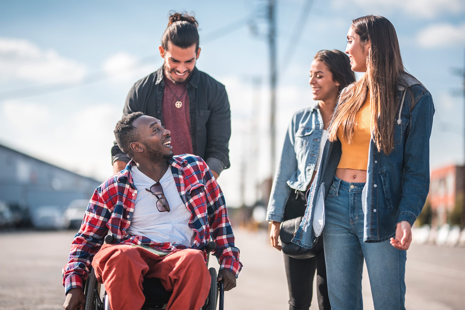 Four friends walk together outside on a sunny day. One man is in a wheelchair, smiling at his mates; another friend is pushing the wheelchair. Everyone appears happy and engaged in conversation.
