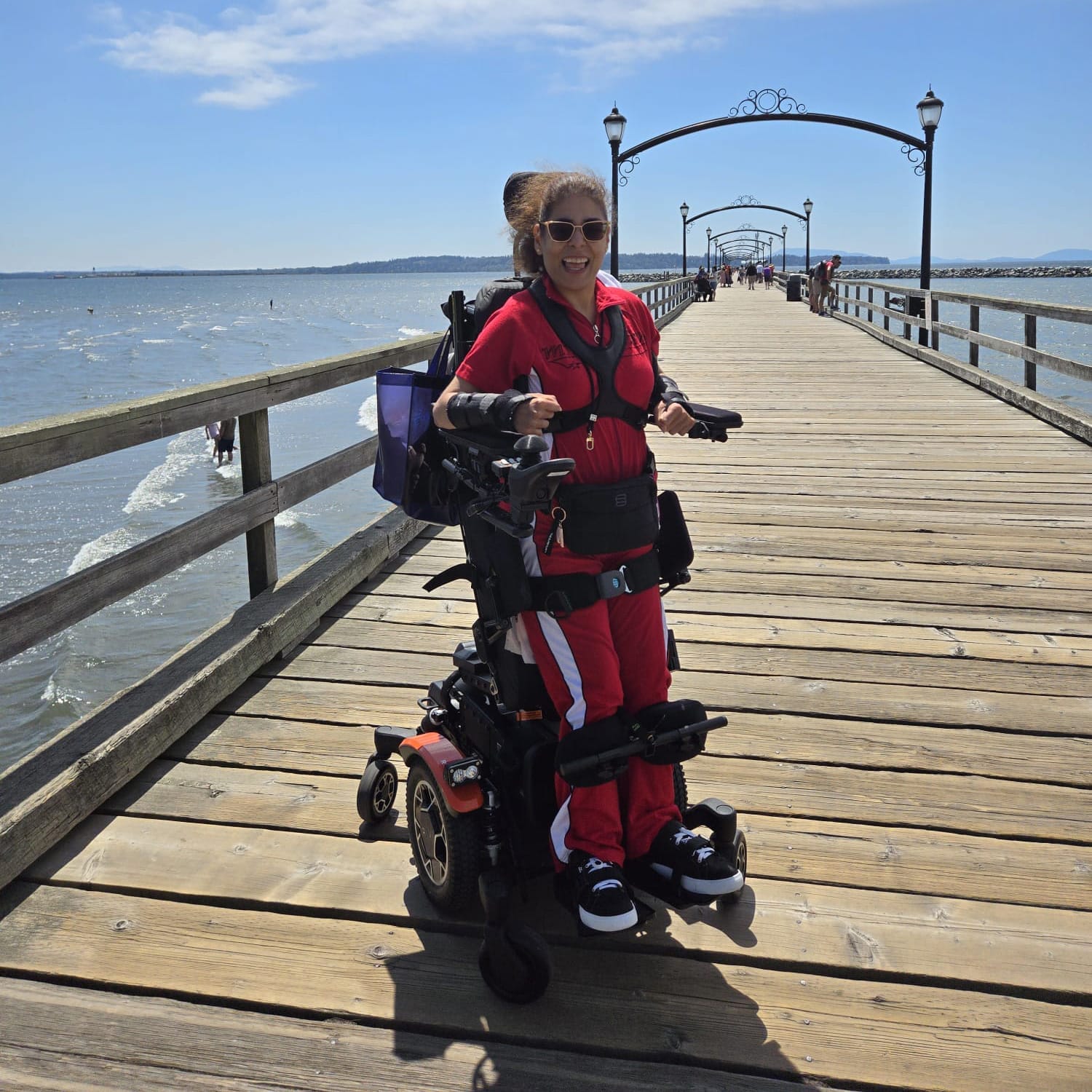 A woman in a red outfit smiles while using a powered wheelchair on a wooden pier extending over the water, with blue sky and distant hills in the background.