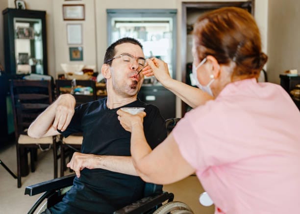A woman in a pink shirt feeds a man in a wheelchair with a spoon. The man, who appears to have a disability, is wearing a black shirt and glasses. They are indoors, and the woman is wearing a face mask.