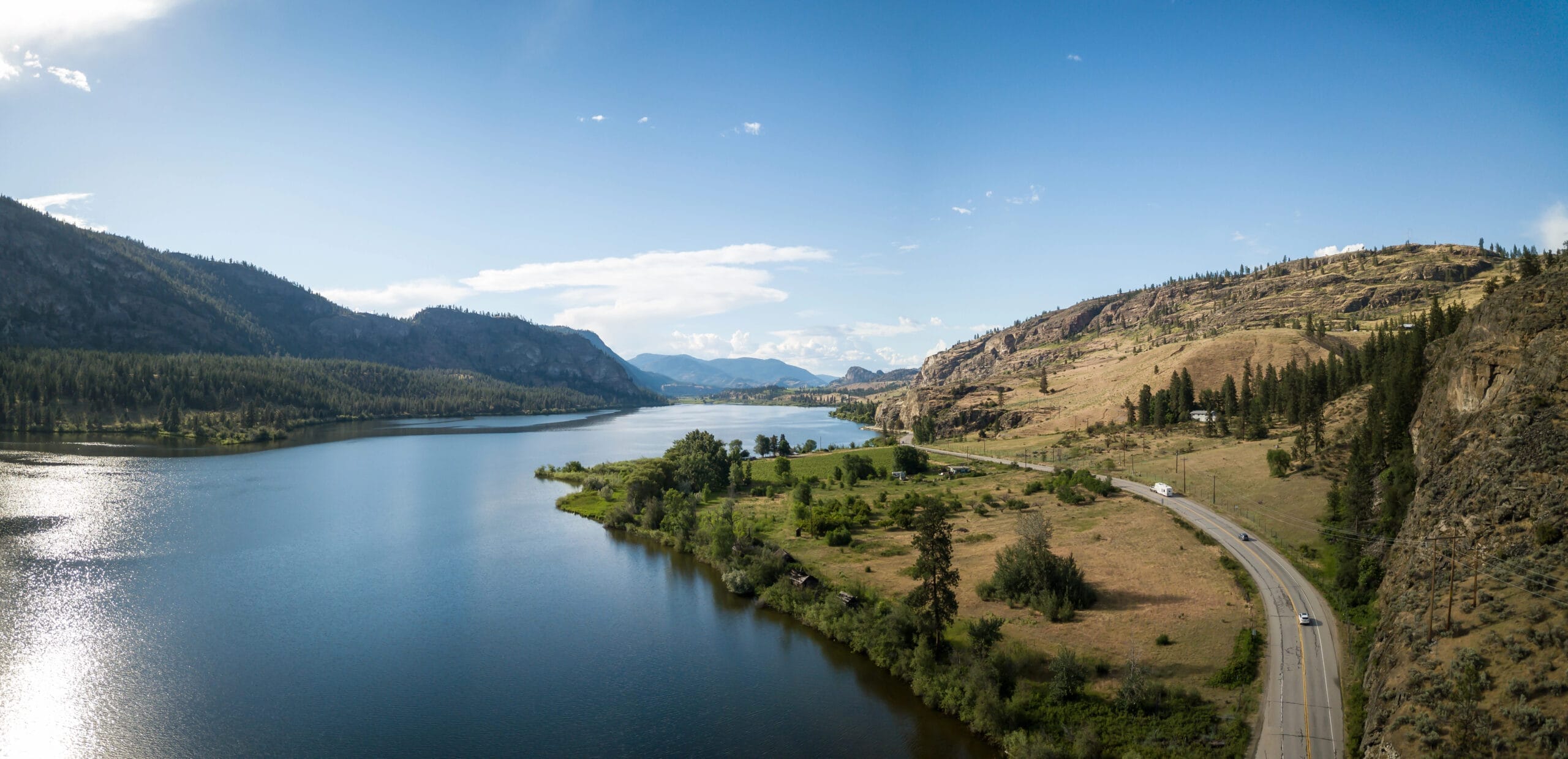 A wide river curves through a green valley bordered by forested hills and rocky slopes under a clear blue sky, with a road winding alongside the water.