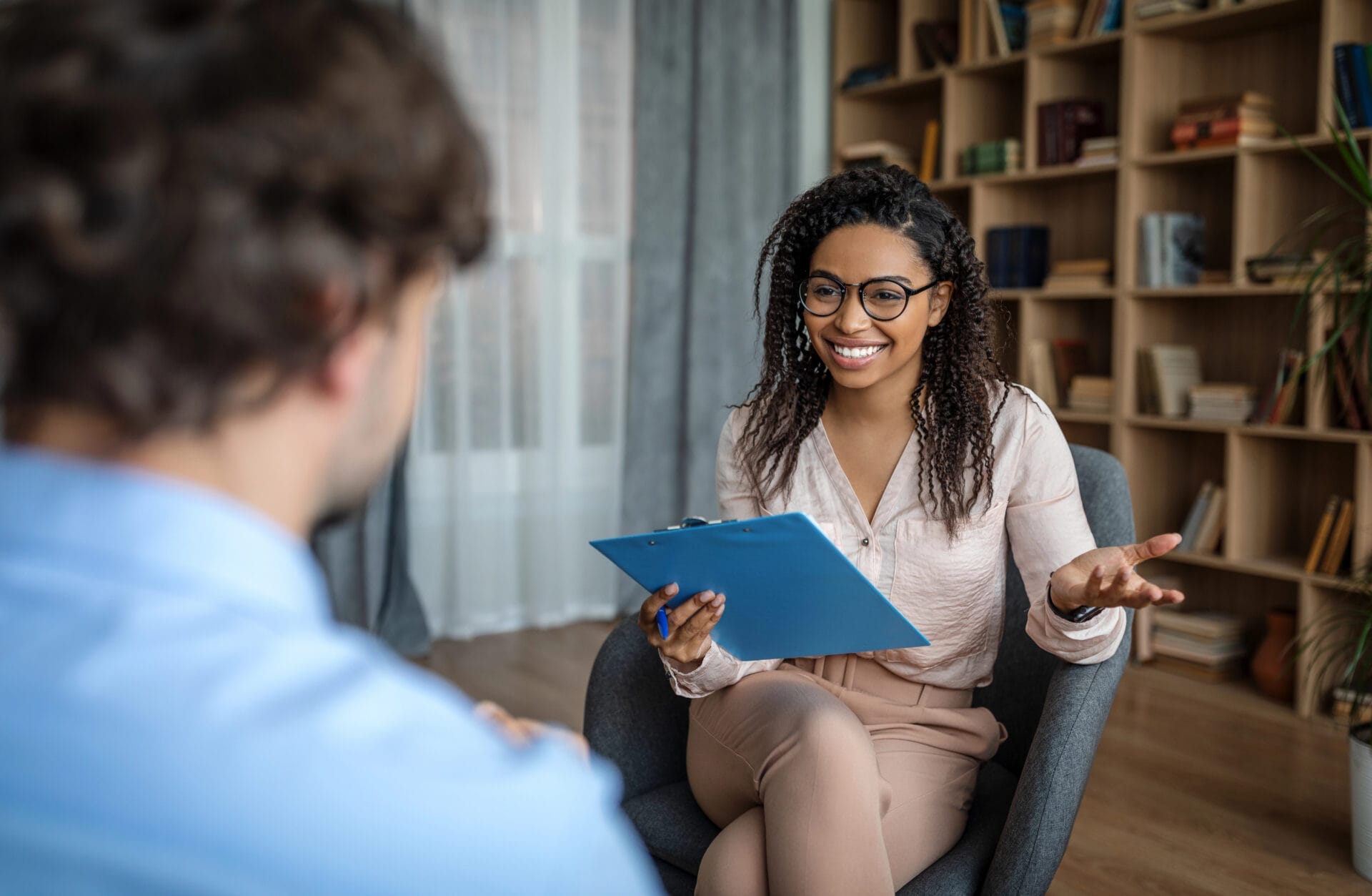 A woman with glasses holds a clipboard and smiles whilst talking to a seated man in an office with bookshelves in the background.