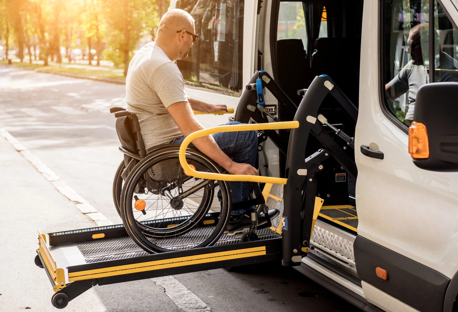 A man in a wheelchair uses a hydraulic lift to board a white accessible van on a sunny day, highlighting accessible public transport.