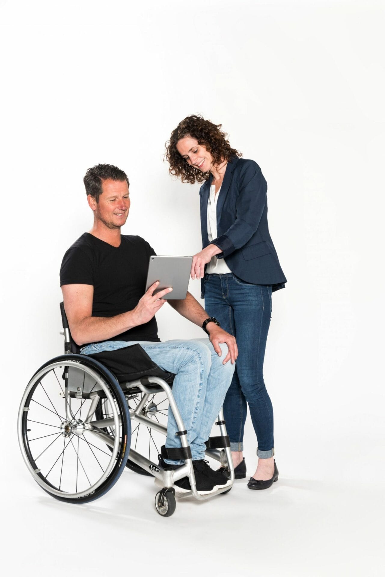A man in a wheelchair holds a tablet while a woman standing next to him points at the screen. Both are smiling and appear to be discussing something on the tablet.