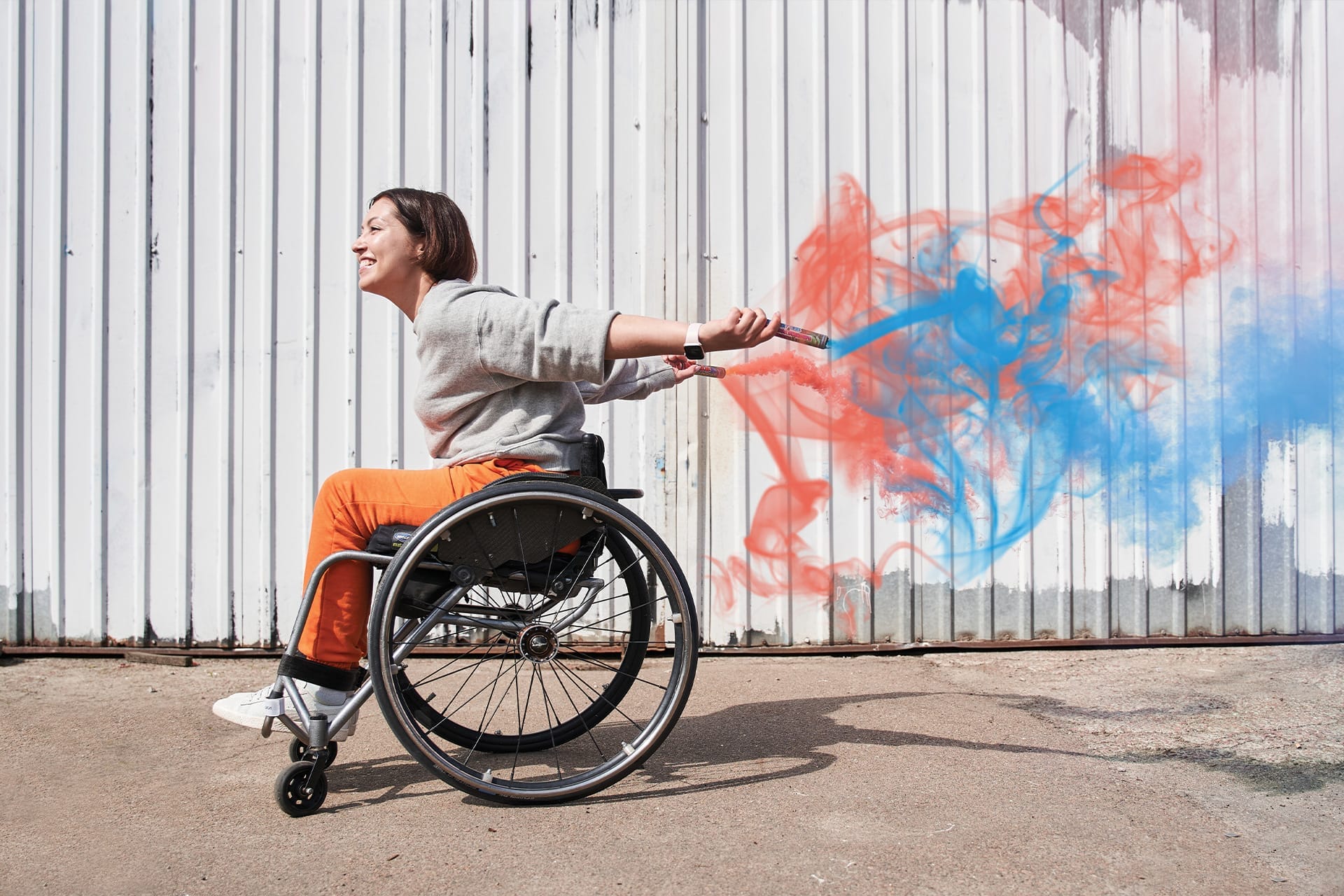 A woman in a wheelchair smiles joyfully, arms outstretched, holding sticks that release colourful red and blue smoke against a corrugated metal background.