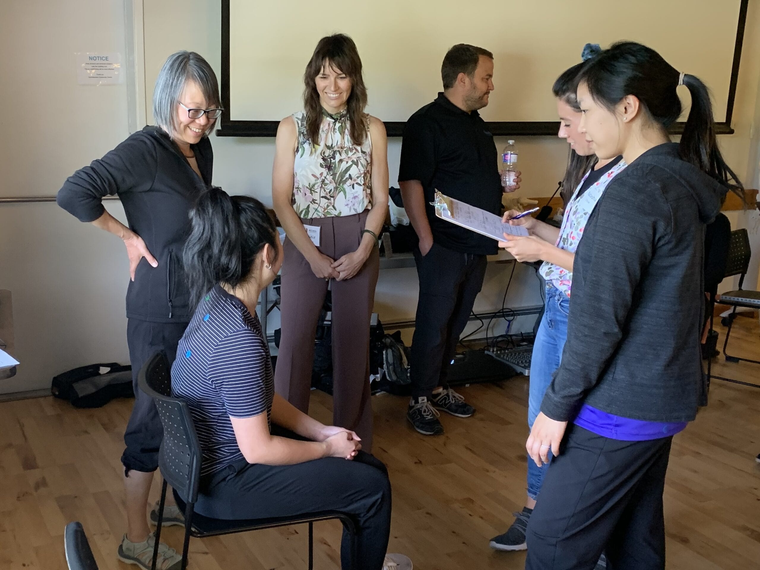 Five people are gathered in a room. One woman sits on a chair whilst the other four stand around her, talking and smiling. Two standing women hold a clipboard and water bottle. The atmosphere appears friendly and collaborative.