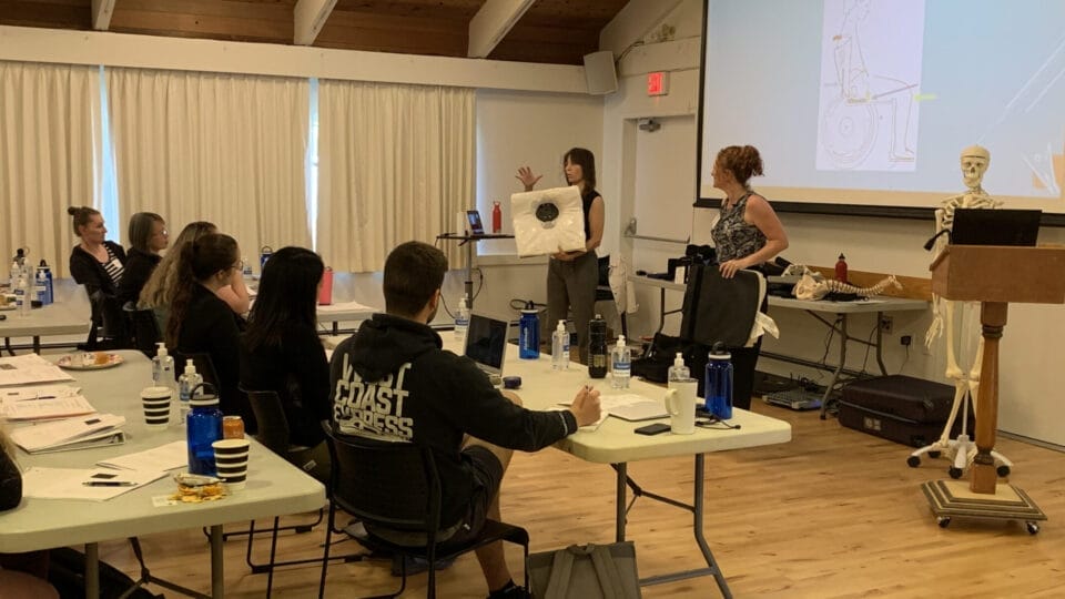 Danielle Rae is at the front of a classroom holding up a diagram, while Lindsay Alford assists. Several people are seated at tables, listening and taking notes. A skeleton model and a projector screen are also visible.